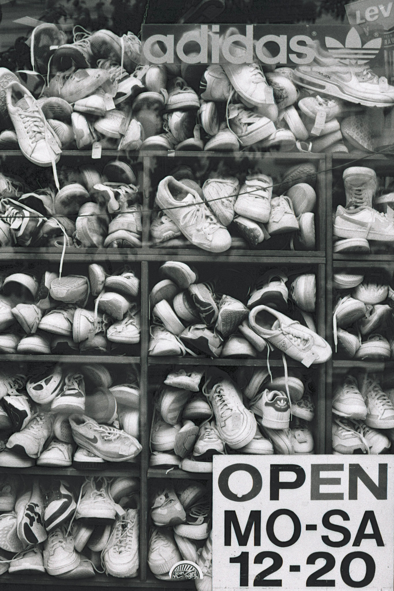 A shop window stacked with worn white sneakers, with a sign reading "Open Mo–Sa 12–20".